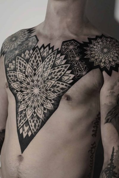 7sinstattoo.live Osoba prezentuje misterny i rozległy czarno-szary tatuaż mandali, który rozciąga się na klatce piersiowej i ramionach, z dodatkowymi mniejszymi tatuażami zdobiącymi jej ramię. Oświetlenie podkreśla szczegółowy projekt i cieniowanie widocznego tatuażu na klatce piersiowej, umiejętnie wykonanego w tatuaż Poznań, jak uchwycono w produkcie DSC04850. Najlepsze studio tatuażu Seven Sins Tattoo