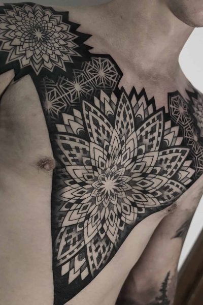 7sinstattoo.live Osoba z misternymi czarno-szarymi geometrycznymi tatuażami mandali DSC04864 na klatce piersiowej i ramieniu, wykonanymi w najlepszym studio tatuażu w Poznaniu. Tatuaże składają się z warstwowych, symetrycznych wzorów o różnych kształtach i cieniowaniu, pokrywających jedno ramię i rozciągających się na górną część klatki piersiowej. Najlepsze studio tatuażu Seven Sins Tattoo