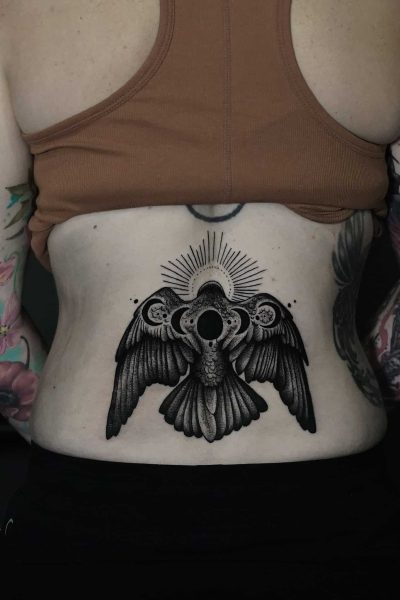 7sinstattoo.live Osoba oznaczona jako DSC05694 ma tatuaż na plecach przedstawiający szczegółowego, ciemno cieniowanego ptaka z rozpostartymi skrzydłami, okrągłym wzorem przypominającym słońce nad głową i misterną kropką. Ma na sobie brązowy krótki top i ciemne spodnie, odsłaniając kolorowe tatuaże na ramionach wykonane w najlepszym studiu tatuażu Poznań. Najlepsze studio tatuażu Seven Sins Tattoo