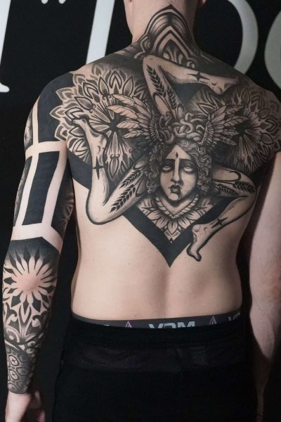 7sinstattoo.live Na plecach osoby znajduje się szczegółowy czarno-szary tatuaż z geometrycznym wzorem i postacią z misternymi skrzydłami oraz ozdobnym nakryciem głowy, stworzony w najlepszym studio tatuażu w Poznaniu. Osoba ta ma również abstrakcyjne i kwiatowe tatuaże na prawym ramieniu. Więcej szczegółów można znaleźć w nazwie produktu: DSC05897. Najlepsze studio tatuażu Seven Sins Tattoo