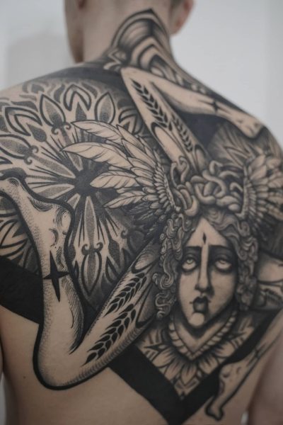 7sinstattoo.live Na plecach osoby znajduje się DSC07467, misterny czarno-szary tatuaż. Skomplikowany projekt przedstawia centralną postać z szczegółową twarzą i włosami, otoczoną symetrycznymi, abstrakcyjnymi i geometrycznymi wzorami, które rozciągają się na górnej części pleców i ramionach. To arcydzieło zostało wykonane w jednym z najlepszych studiów tatuażu w Poznaniu. Najlepsze studio tatuażu Seven Sins Tattoo
