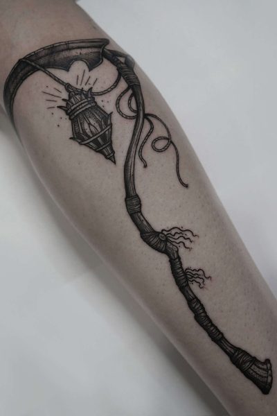 7sinstattoo.live Tatuaż DSC07641 na ramieniu przedstawia szczegółowo odwzorowaną czarno-szarą latarnię uliczną zawieszoną na zakrzywionym, zwiniętym słupie z małym toporem na końcu. Lampa emituje świecące promienie światła. Słup zdobią misterne wiązania i wstążki, mistrzowsko wykonane przez najlepsze studio tatuażu w Poznaniu. Najlepsze studio tatuażu Seven Sins Tattoo