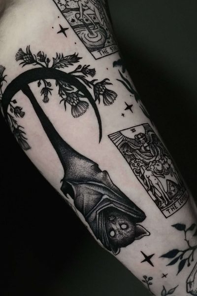 7sinstattoo.live Tatuaż na ramieniu wykonany w najlepszym studio tatuażu Poznań zawiera misterne wzory wykonane czarnym tuszem, w tym dużego nietoperza wiszącego do góry nogami, elementy kwiatowe i dwie karty tarota oznaczone jako „The Sun” i „The Lovers”. Obszar otaczający obejmuje liście i detale związane z naturą. To oszałamiające dzieło sztuki zostało uchwycone w produkcie DSC07762. Najlepsze studio tatuażu Seven Sins Tattoo