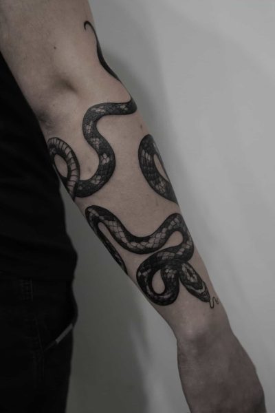 7sinstattoo.live Zdjęcie z bliska, zidentyfikowane jako DSC09448, przedstawia przedramię osoby z misternym tatuażem czarnego węża, który wije się od nadgarstka w kierunku łokcia. Ta szczegółowa grafika została wykonana w jednym z najlepszych studiów tatuażu w Poznaniu. Osoba ma jasną skórę i nosi czarną koszulę z krótkim rękawem na jednolitym, jasnym tle. Najlepsze studio tatuażu Seven Sins Tattoo