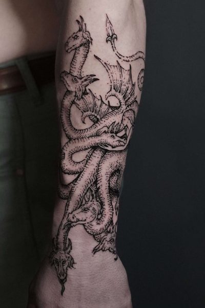 7sinstattoo.live Zbliżenie na przedramię osoby, na którym widoczny jest skomplikowany tatuaż wielogłowego smoka, wykonany przez utalentowanego artystę z najlepszego studia tatuażu w Poznaniu. Szczegółowa praca liniowa ukazuje głowy smoków warczące i splatające się wzdłuż przedramienia. Osoba, zidentyfikowana jako DSC09812, ma na sobie zielone spodnie. Najlepsze studio tatuażu Seven Sins Tattoo