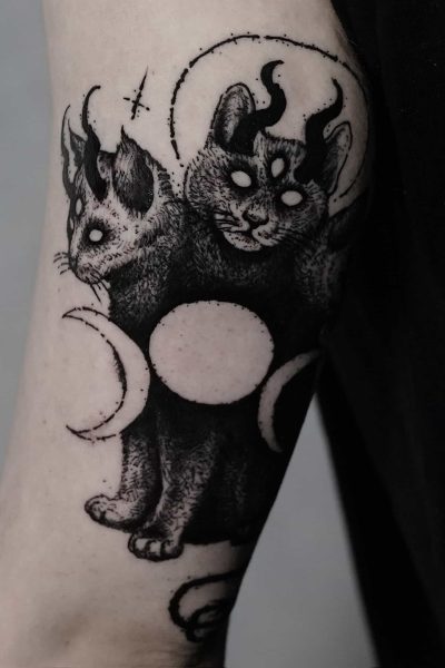 7sinstattoo.live Oszałamiający tatuaż zatytułowany „DSC09934”, stworzony w najlepszym studio tatuażu Poznań, przedstawia dwugłowego kota z trzema oczami i rogami. Obie głowy mistycznego stworzenia mają świecące oczy i są otoczone elementami niebieskimi, w tym fazami księżyca. Ta czarno-biała grafika zdobi ramię osoby na ciemnym tle. Najlepsze studio tatuażu Seven Sins Tattoo