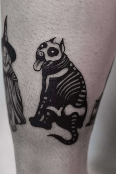 7sinstattoo.live Tatuaż na nodze osoby przedstawiający czarno-białego stylizowanego psa o szkieletowych rysach i wystającym języku, obok częściowego obrazu postaci w szacie i kapeluszu. Projekt utrzymuje mroczny, kapryśny motyw i został wykonany przez artystę z najlepszego studia tatuażu Poznań. Ta praca jest identyfikowana jako DSC09947. Najlepsze studio tatuażu Seven Sins Tattoo