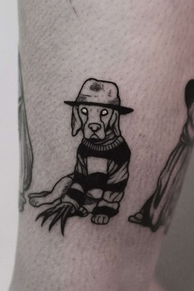 7sinstattoo.live Czarno-biały tatuaż, wykonany w studio tatuaży Poznań, przedstawia psa w szerokim kapeluszu i pasiastym swetrze, przypominającym postać z horroru. Pies ma pazury w rękawiczkach na jednej łapie, co dodaje niepokojącego motywu, i siedzi na jasnym ramieniu osoby. Tatuaż jest skatalogowany jako produkt DSC09972. Najlepsze studio tatuażu Seven Sins Tattoo