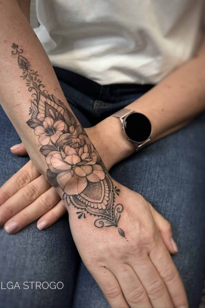 7sinstattoo.live Przedramię prezentujące IMG-5630, misterny czarno-szary kwiatowy tatuaż mandali rozciągający się na dłoń i nadgarstek. Osoba jest ubrana w biały top, niebieskie dżinsy i srebrny smartwatch. Ten misterny tatuaż, wykonany w Najlepszym Studio Tatuażu Poznań, zawiera szczegółowe kwiaty i ozdobne wzory z tekstem „OLGA STROGO. Najlepsze studio tatuażu Seven Sins Tattoo