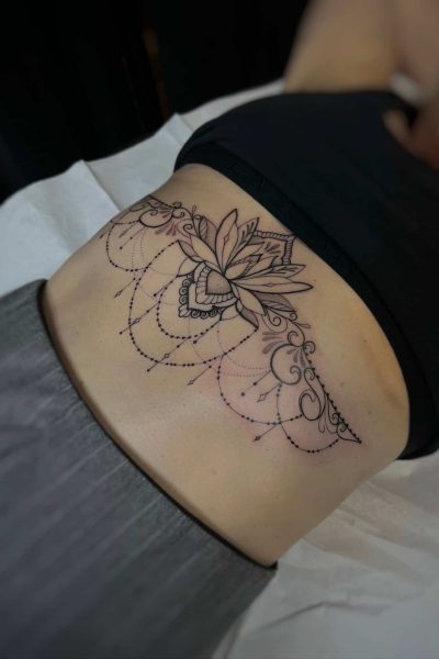 7sinstattoo.live W IMG-5736 szczegółowe zbliżenie ukazuje dolną część pleców osoby ozdobioną misternym tatuażem z cenionego studia tatuażu Poznań. Tatuaż przedstawia widoczny kwiat lotosu w centrum, otoczony misternymi, symetrycznymi wzorami i precyzyjnymi kropkami, które rozciągają się na dolnej części pleców. Osoba jest umieszczona na białym prześcieradle. Najlepsze studio tatuażu Seven Sins Tattoo