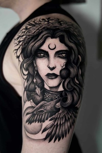 7sinstattoo.live Osoba ze szczegółowym czarno-szarym tatuażem na górnej części ramienia. Tatuaż „fggdh” Poznań przedstawia kobietę z długimi, falującymi włosami, półksiężycem na czole i dużym ptakiem, prawdopodobnie krukiem, na piersi — z misternym cieniowaniem i elementami wzornictwa. Najlepsze studio tatuażu Seven Sins Tattoo