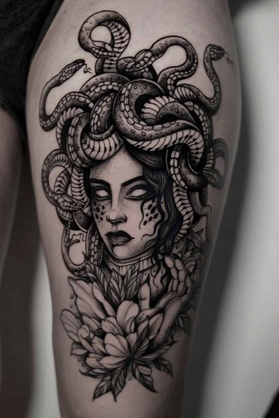 7sinstattoo.live Na udzie osoby znajduje się szczegółowy tatuaż z hjkl, studia tatuaży w Poznaniu, przedstawiający mityczną głowę Meduzy z wężami zamiast włosów. Wzór obejmuje skomplikowane wzory węży splecionych wokół jej głowy i kwiatów kwitnących poniżej jej szyi, tworząc uderzającą kompozycję w czerni i szarości. Najlepsze studio tatuażu Seven Sins Tattoo