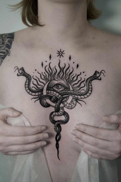 7sinstattoo.live Osoba z dużym tatuażem na klatce piersiowej przedstawiającym oko otoczone płomieniami i splecionymi wężami trzyma lekką, półprzezroczystą tkaninę, częściowo zakrywającą dolną część tatuażu. Czarno-szary tatuaż kwiatowy, stworzony przez studio tatuażu Poznań, zdobi jej górną część lewego ramienia. Ten element nosi nazwę jsnhj. Najlepsze studio tatuażu Seven Sins Tattoo