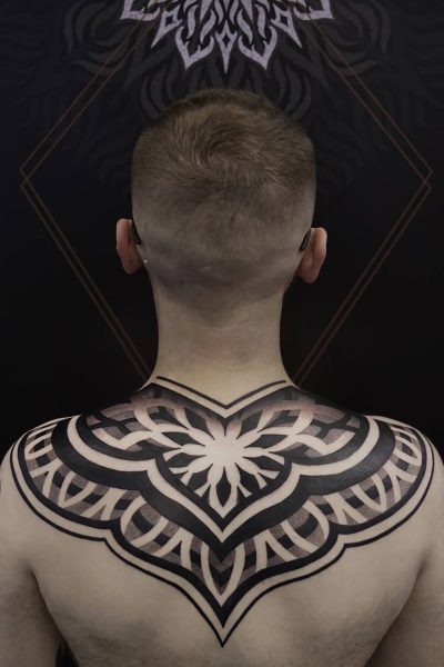7sinstattoo.live Osoba z fryzurą na jeża jest przedstawiona od tyłu, odsłaniając skomplikowany i symetryczny czarny tatuaż, który rozciąga się na górnej części pleców i ramionach. Projekt prezentuje geometryczne wzory przeplatane elementami kwiatowymi. Ciemne tło, ozdobione subtelnymi geometrycznymi kształtami, podkreśla kunszt poznańskiego eksperta tatuażu przy użyciu produktu x49. Najlepsze studio tatuażu Seven Sins Tattoo