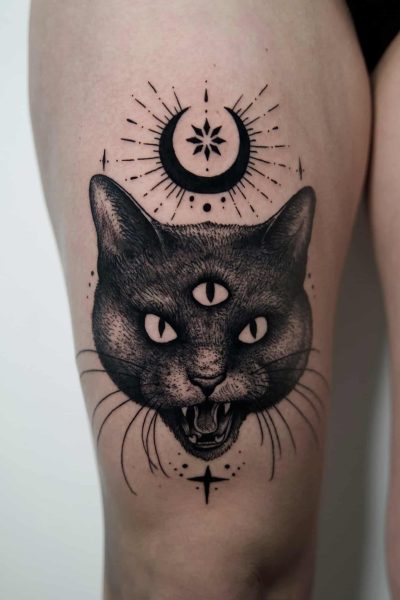7sinstattoo.live Czarno-szary tatuaż Poznań na udzie człowieka przedstawia głowę kota z trzema oczami, otwartą paszczą pokazującą zęby i szczegółowe wąsy. Nad kotem znajduje się półksiężyc, gwiazda i dodatkowe elementy dekoracyjne. Odwiedź najlepsze studio tatuażu Poznań, aby uzyskać wyjątkową sztukę tatuażu z wzorami takimi jak Копия DSC03086оьоо. Najlepsze studio tatuażu Seven Sins Tattoo