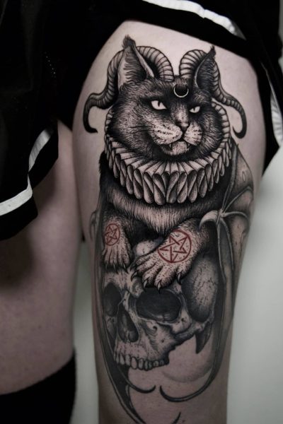 7sinstattoo.live Szczegółowy tatuaż, uchwycony na zdjęciu produktu „Копия DSC09932” przez utalentowanego tatuażystę z Poznania, przedstawia kota z rogami barana i misterną obrożą, spoczywającego na ludzkiej czaszce. Na łapach kota znajdują się dekoracyjne symbole. Tatuaż jest umieszczony na udzie osoby, z widocznym brzegiem jej czarnego ubrania. Najlepsze studio tatuażu Seven Sins Tattoo