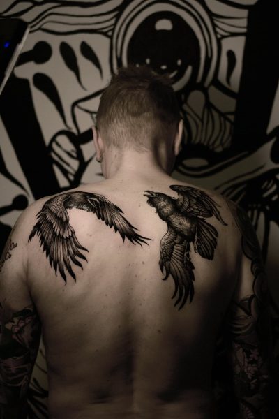 7sinstattoo.live Osoba z krótkimi włosami i widocznymi tatuażami na ramionach jest widziana od tyłu, prezentując dwa duże tatuaże ptaków na górnej części pleców. Tło przedstawia czarno-białą abstrakcyjną sztukę muralową. Obraz ma słabe, dramatyczne oświetlenie, przedstawiające wykwintną pracę najlepszego studia tatuażu Poznań. Najlepsze studio tatuażu Seven Sins Tattoo