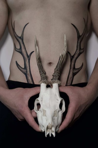 7sinstattoo.live Osoba bez koszulki trzyma w rękach czaszkę jelenia, wyrównując jej poroże z misternymi czarnymi tatuażami poroża na brzuchu, tworząc iluzję, w której tatuaże płynnie rozciągają się od czaszki. Mają na sobie ciemne spodnie i stoją w renomowanym studiu tatuażu w Poznaniu. Ta artystyczna scena pięknie uosabia unikalny styl прррр. Najlepsze studio tatuażu Seven Sins Tattoo